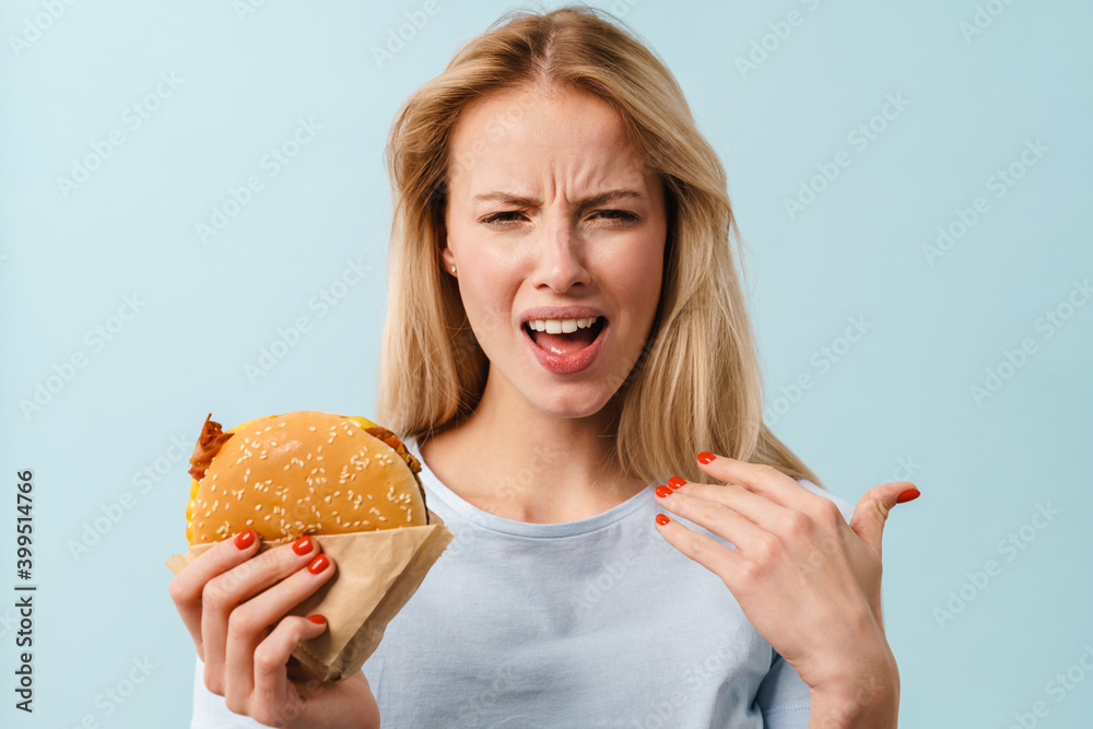 Displeased beautiful girl grimacing while posing with hamburger Displeased beautiful girl grimacing while posing with hamburger