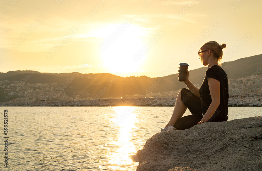 Woman with tumbler cup sitting on a rock of cliff with sea and cityscape on background during sunset, copy space