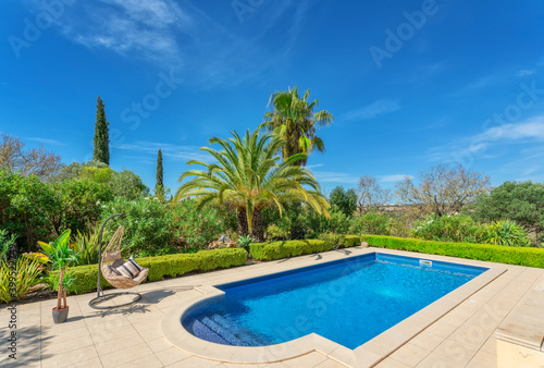 Luxurious pool in the garden of a private villa, hanging chair with pillows for leisure tourists, in summer. Portugal, Algarve. © sergojpg