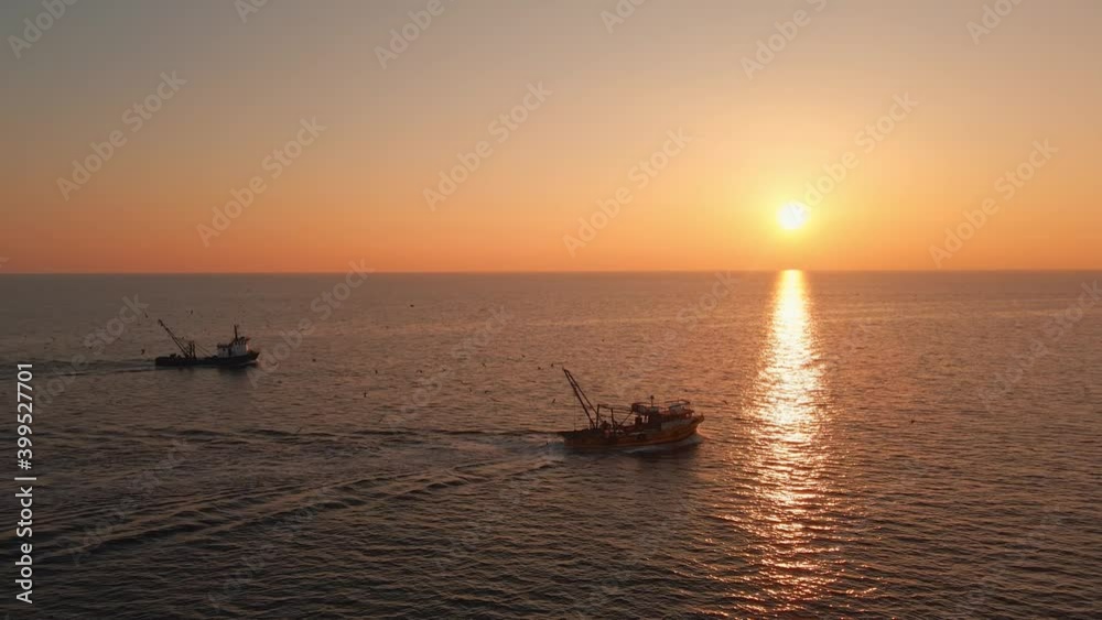 Fishing Boat with Large Catch Fish Swirling Flock Gulls Aerial View Drone Sunset Winter. Small Ship Floats on Sea Surface Golden Sky Leaving a Path of Sea Foam Water. Seagulls. Bright Disk of Sun