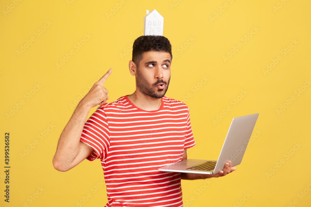 Funny bearded man in striped t-shirt pointing at paper house on his head standing and holding laptop, working from home, freelance, teleworking. Indoor studio shot isolated on yellow background Funny bearded man in striped t-shirt pointing at paper house on his head standing and holding laptop, working from home, freelance, teleworking. Indoor studio shot isolated on yellow background