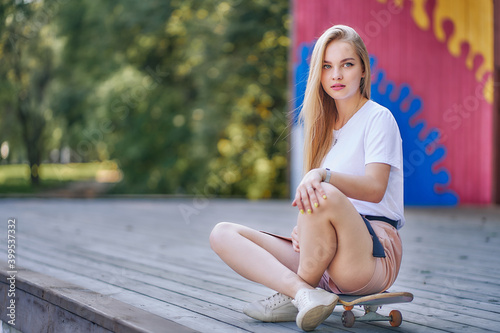 young woman sits cross-legged with a skateboard on the summer terrace of the park.