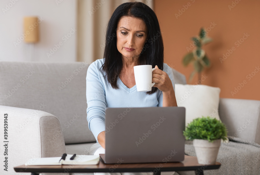 Mature woman sitting on sofa drinking coffe working on computer Mature woman sitting on sofa drinking coffe working on computer