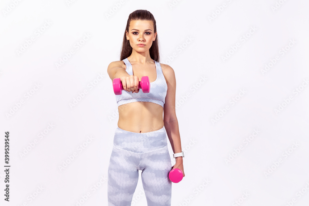 Confident assertive sports woman in white top and tights doing sports exercises holding dumbbells in hands, pumping up muscles, developing strength. Indoor studio shot isolated on gray background Confident assertive sports woman in white top and tights doing sports exercises holding dumbbells in hands, pumping up muscles, developing strength. Indoor studio shot isolated on gray background