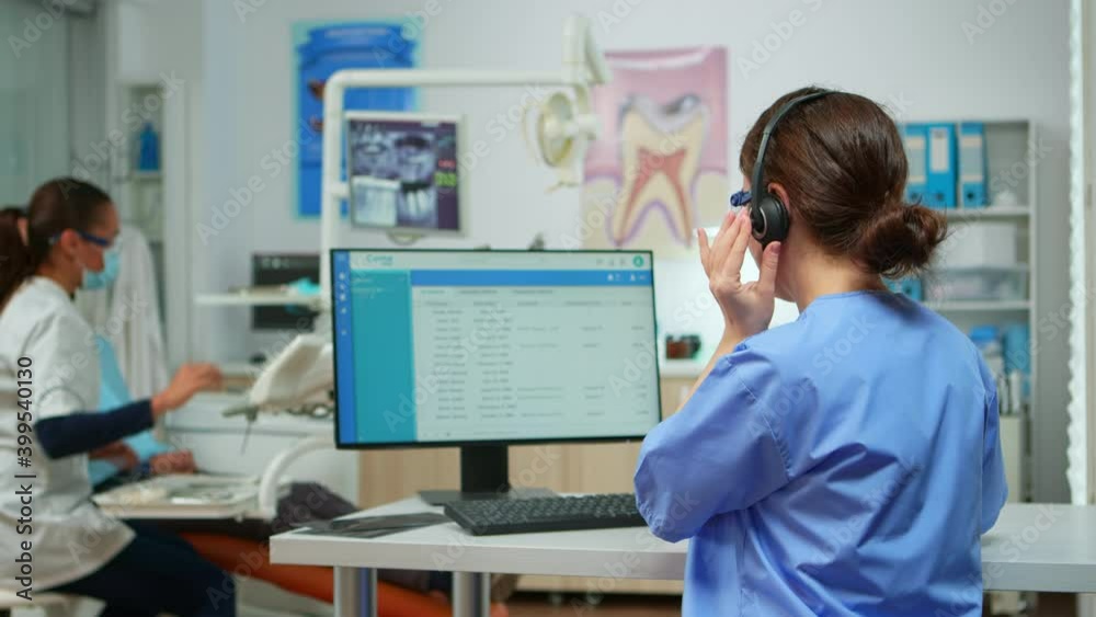 Dentist assistant making appointments using headset sitting in front on computer while doctor is working with patient in background examining teeth problem. Nurse taking notes in stomatological office Dentist assistant making appointments using headset sitting in front on computer while doctor is working with patient in background examining teeth problem. Nurse taking notes in stomatological office