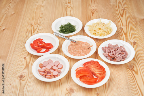 Seven white plates with chopped pizza ingredients on a light wooden background. Close-up. View from above.