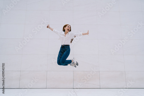 Excited young woman in casual outfit jumping against gray modern building wall