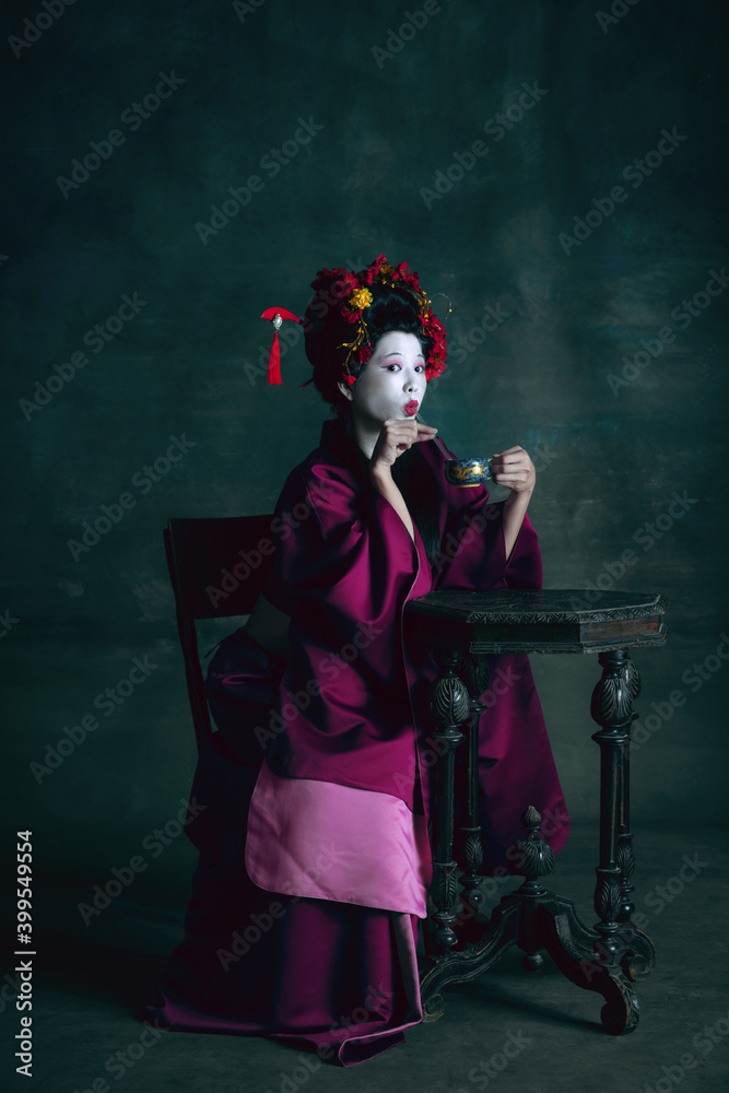 Tea drinking. Young japanese woman as geisha isolated on dark green background. Retro style, comparison of eras concept. Beautiful female model like bright historical character, old-fashioned.