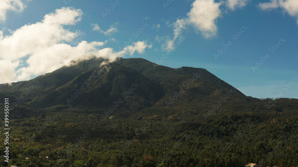 Fototapeta premium Mountains covered rainforest, trees and blue sky with clouds, aerial view. Camiguin, Philippines. Mountain landscape on tropical island with mountain peaks covered with forest. Slopes of mountains