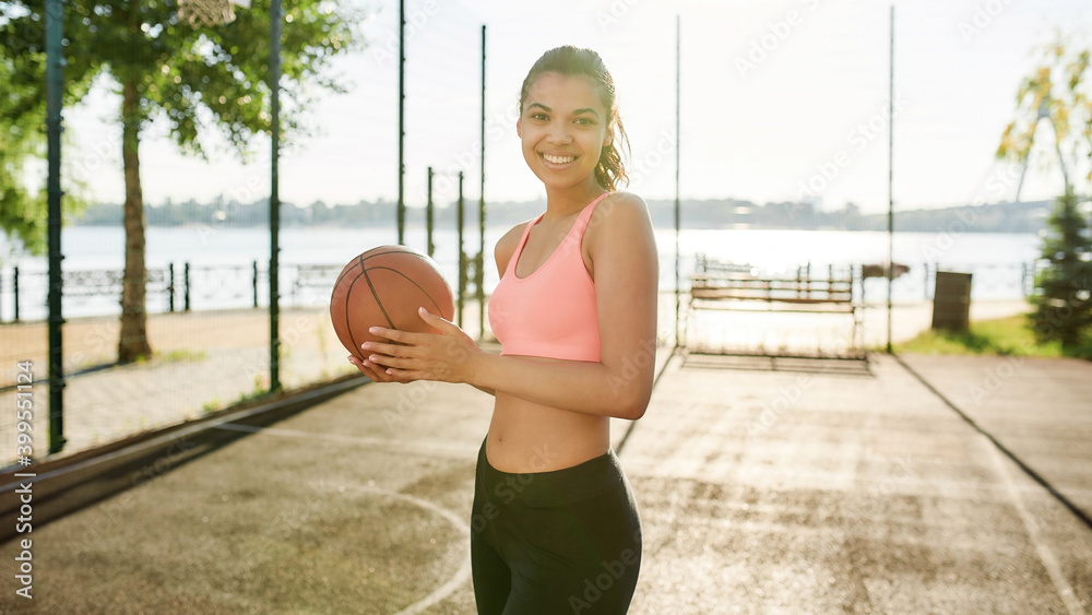Beautiful young mixed race woman in sportswear smiling at camera while posing with basketball outdoors on a sunny day Beautiful young mixed race woman in sportswear smiling at camera while posing with basketball outdoors on a sunny day