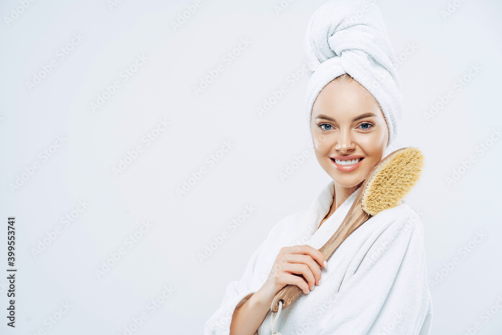 Sideways shot of beautiful young European woman with charming smile, poses with bath item, uses brush to massage body, poses against white background, blank space for your promotional content Sideways shot of beautiful young European woman with charming smile, poses with bath item, uses brush to massage body, poses against white background, blank space for your promotional content