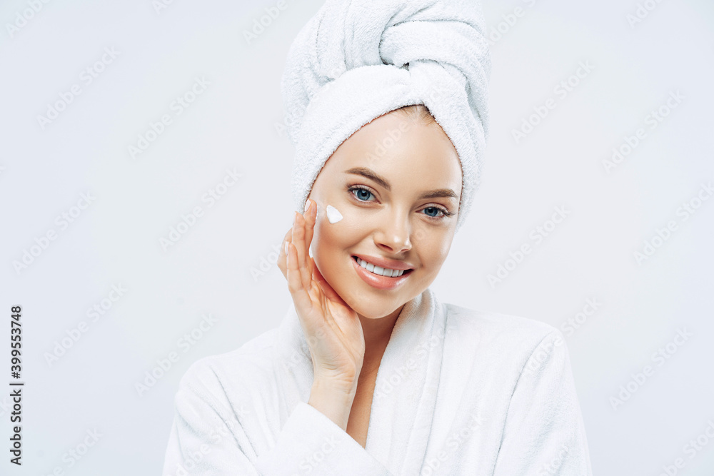Charming relaxed beauty woman applies face cream, cares about complexion, touches cheek with hand, smiles gently at camera, dressed in bath robe, wrapped towel on washed hair, isolated on white Charming relaxed beauty woman applies face cream, cares about complexion, touches cheek with hand, smiles gently at camera, dressed in bath robe, wrapped towel on washed hair, isolated on white