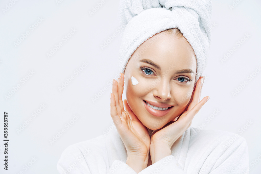 Close up portrait of cheerful female with natural beauty, touches cheeks gently, applies face cream for soft skin, wears bath towel on head, dressed in robe, stands indoor. Beauty treatment, lifestyle Close up portrait of cheerful female with natural beauty, touches cheeks gently, applies face cream for soft skin, wears bath towel on head, dressed in robe, stands indoor. Beauty treatment, lifestyle