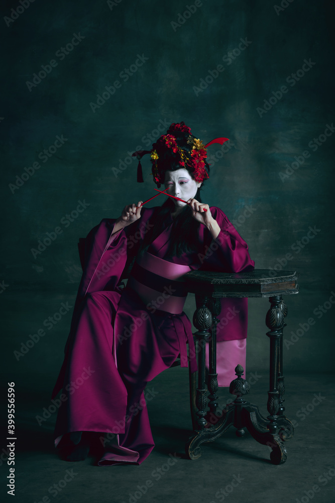 Fototapeta premium Playing with chopsticks. Young japanese woman as geisha isolated on dark green background. Retro style, comparison of eras concept. Beautiful female model like bright historical character, old