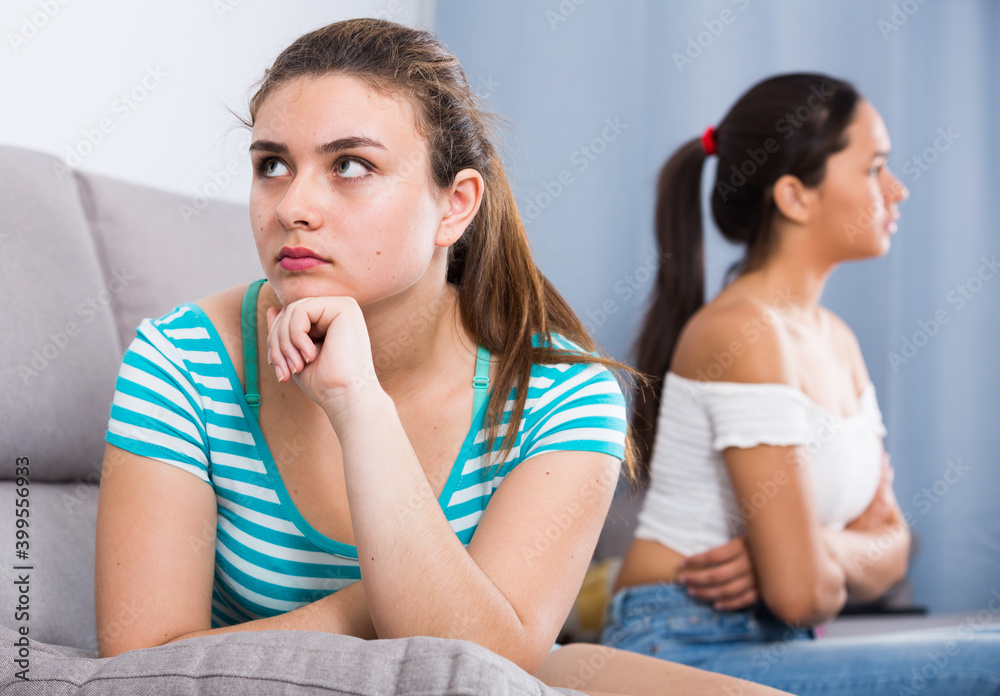 Two angry quarreled teenage girls sitting apart on couch at home