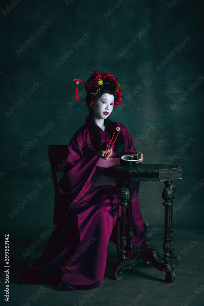 Eating sushi. Young japanese woman as geisha isolated on dark green background. Retro style, comparison of eras concept. Beautiful female model like bright historical character, old-fashioned.
