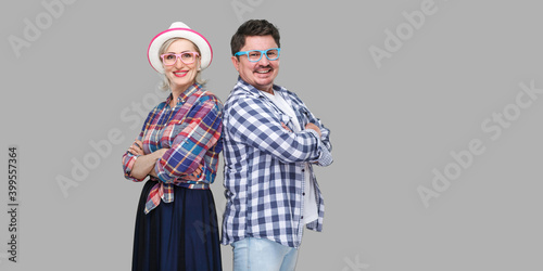 Couple of friends, adult man and woman in casual checkered shirt standing together back to back with crossed arms, toothy smile, looking at camera. Indoor, isolated, studio shot, gray background