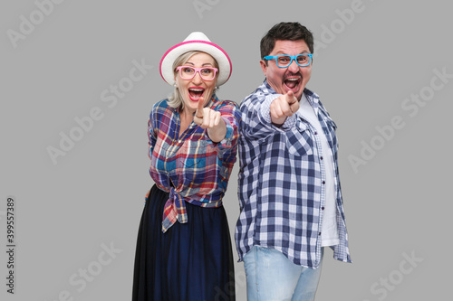 Couple of friends, adult man and woman in casual checkered shirt standing together back to back pointing finger, looking amazed, surprised or laughing. Indoor,isolated,studio shot, gray background