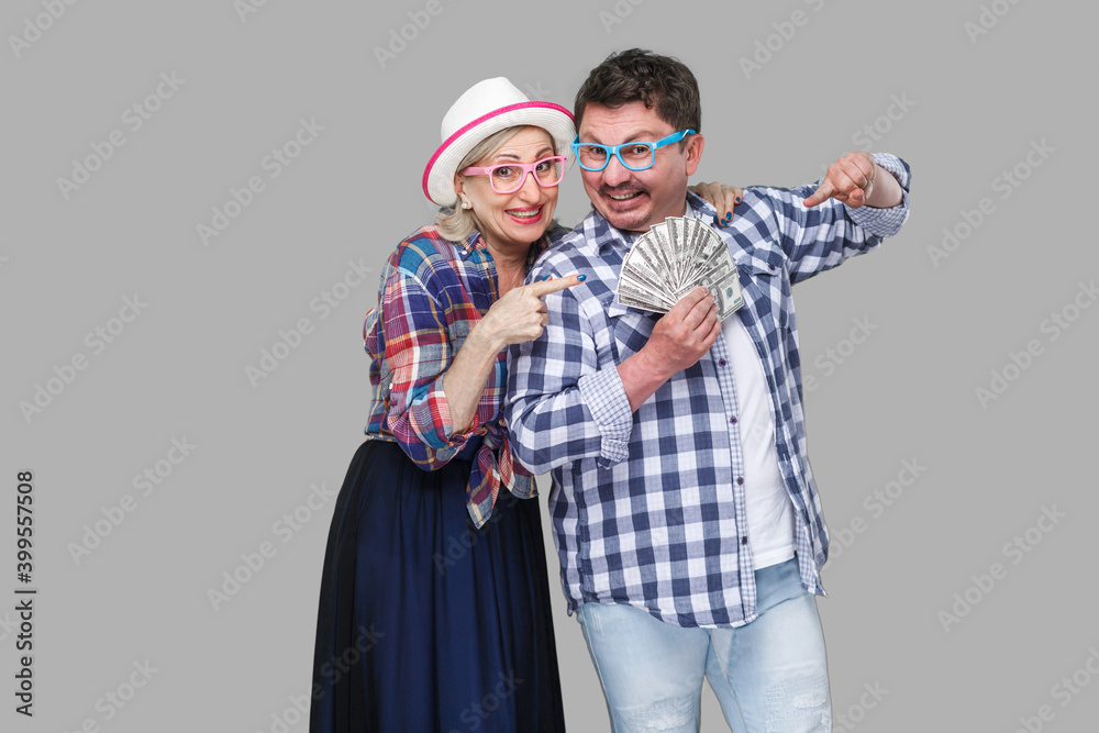 Happy wealthy family, adult man and woman in casual checkered shirt standing pickaback together, holding fan of dollar and pointing finger to money. Indoor,isolated, studio shot, gray background Happy wealthy family, adult man and woman in casual checkered shirt standing pickaback together, holding fan of dollar and pointing finger to money. Indoor,isolated, studio shot, gray background