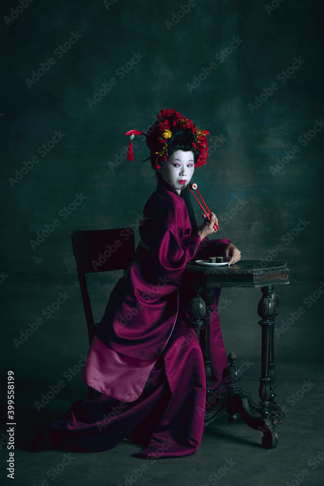 Fototapeta premium Tasty sushi. Young japanese woman as geisha isolated on dark green background. Retro style, comparison of eras concept. Beautiful female model like bright historical character, old-fashioned.