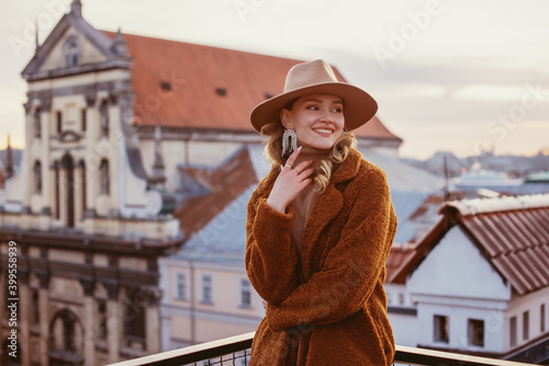 Foto Fashionable happy smiling woman wearing trendy beige hat, rhinestones earrings, brown faux fur coat, posing on balcony with beautiful view on European city