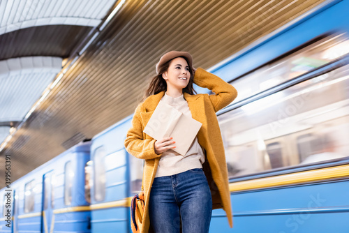 happy woman in autumn outfit holding book and looking at metro train arriving on platform