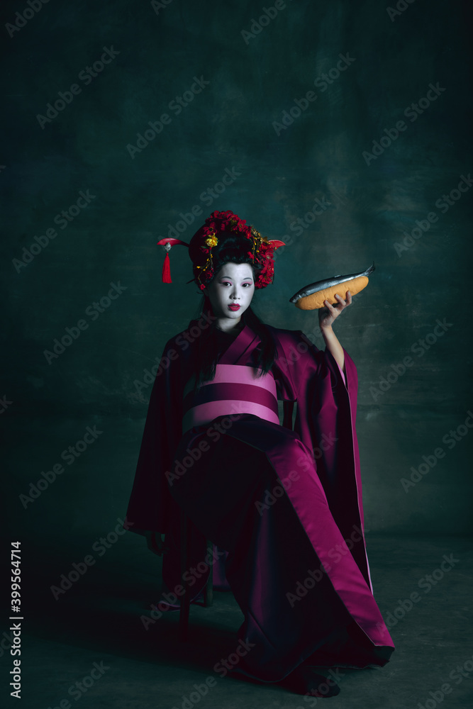 Obraz premium Tasty cuisine. Young japanese woman as geisha isolated on dark green background. Retro style, comparison of eras concept. Beautiful female model like bright historical character, old-fashioned.