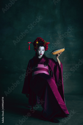 Tasty cuisine. Young japanese woman as geisha isolated on dark green background. Retro style, comparison of eras concept. Beautiful female model like bright historical character, old-fashioned.