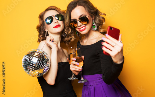 Canvas Print two fashionable young women holding disco ball, drinking champagne and taking a selfie