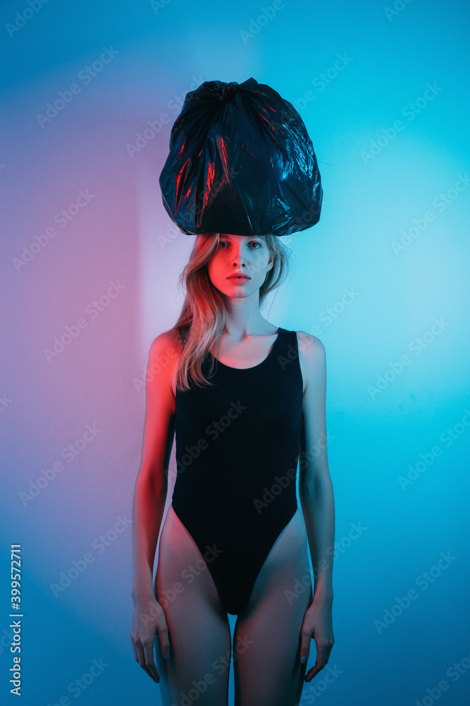 Art portrait. Woman power. Independence challenge. Strong confident brave lady in black bodysuit holding black garbage bag on head in red neon light isolated on blue pink color gradient background.