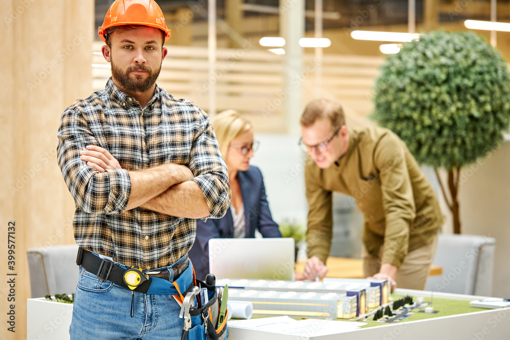 portrait of confident engineer male in safety helmet, young caucasian man posing at camera, they work on planning building project, using layout. colleagues in the background