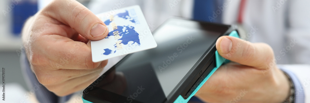 Close-up of adult person holding plastic credit card and machine in hands. Doctor in white uniform and red stethoscope. Online pay and remote transaction concept Close-up of adult person holding plastic credit card and machine in hands. Doctor in white uniform and red stethoscope. Online pay and remote transaction concept