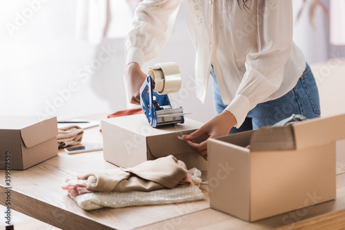 Cropped view of african american businesswoman packing box with adhesive tape near clothes on blurred foreground in showroom photo