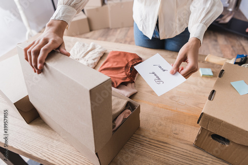 Cropped view of african american owner of showroom holding card with thank you lettering near box with clothes on table