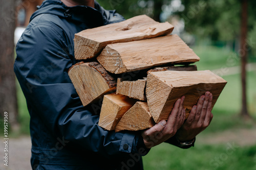 Fototapeta Cropped shot of faceless man in black jacket carries pile of firewood poses against blurred forest background