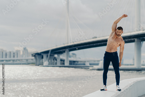 Full length shot of muscles active man does sport exercises outdoor poses near river bridge wears trousers and sneakers has beautiful male torso achieves best shape. Healthy lifestyle concept © VK Studio