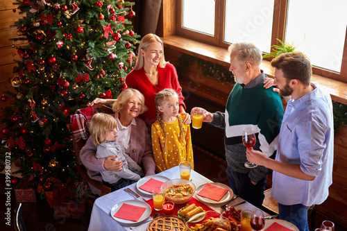 extended family congratulating  talking toasts behind christmas table. elderly man hold glass with beverage in hands and talk