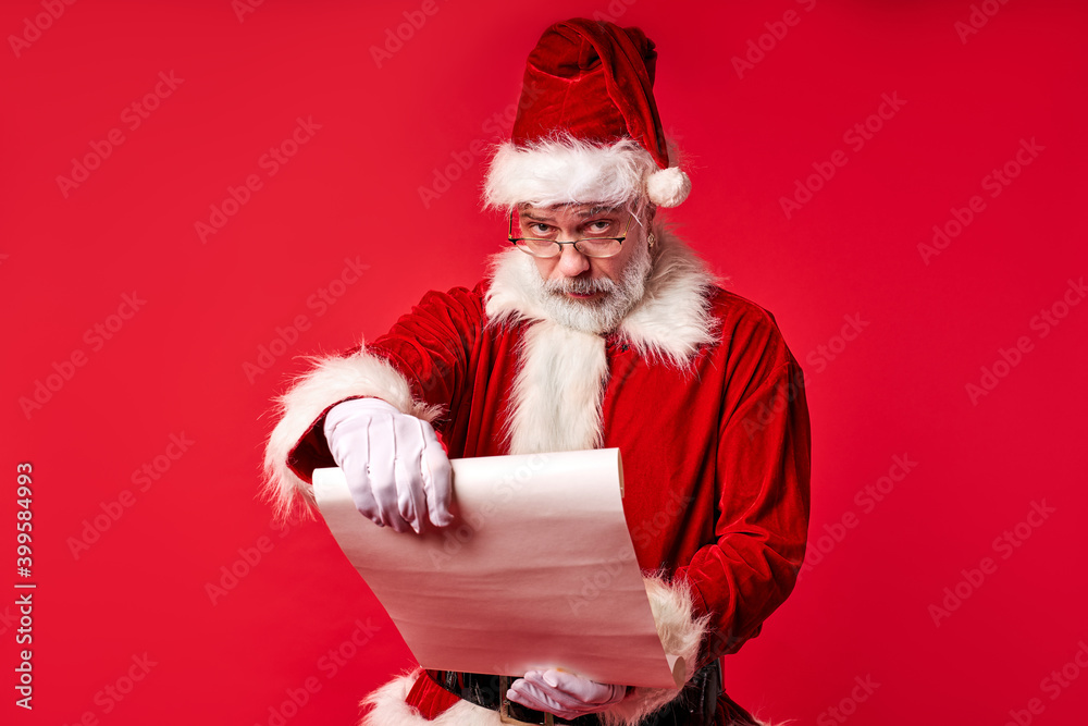 Naklejka premium old bearded Santa Claus reading Merry Christmas wishlist paper roll isolated over red background, senior male in costume with white paper in hands