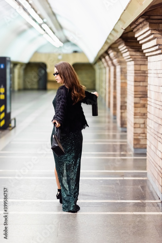 elegant woman in long black dress, faux fur jacket and sunglasses walking with wine bottle along underground station