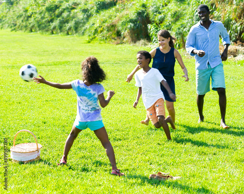 Happy friendly interracial family with preteen children playing ball on green lawn in summer city park