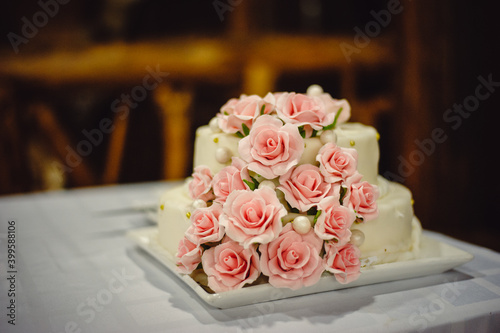 A two-level white wedding cake decorated with pink flowers from a mastic stands on a white tablecloth. Square Mastic Cake. Film noise