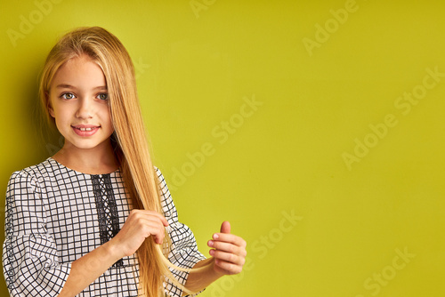 Fotografija portrait of attractive kid girl touching her long straight hair, looking at camera isolated on green studio background, posing