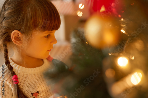 Child dreams near Christmas tree, little girl with in decorated room, side view of child dresses white sweater, kid looking at garlands light with pensive facial expression.