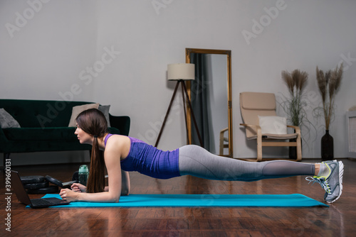 Young woman doing plank in front of laptop while watching online workout session at home in a modern living room