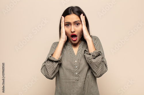 young hispanic woman looking unpleasantly shocked, scared or worried, mouth wide open and covering both ears with hands photo