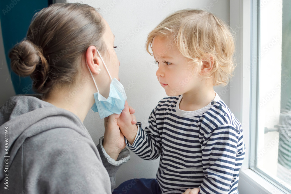 Child in home quarantine taking the medical mask off his mothers face during coronavirus COVID-2019 and flu lock down. Selective focus