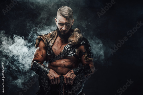 Northern vandal seafarer with muscular build holding huge axe and staring at camera in dark smokey background.