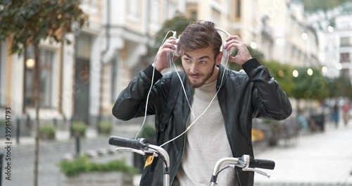 Bike messenger with backpack stands on the street, puts on headphones and listen to music on his smartphone. Cinema 4K 60fps video photo