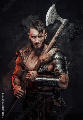Muscular and awesome viking fighter armed with huge axe and dressed in light armour poses in smokey dark background.