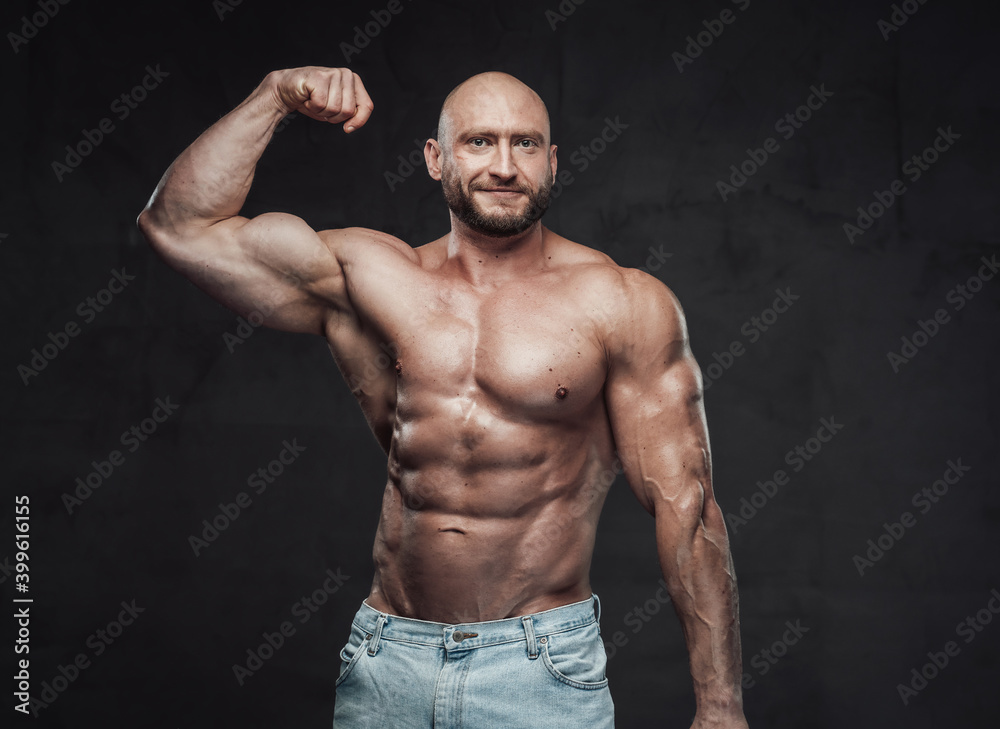 Portrait of a happy and muscular guy with bald head and naked torso posing in dark background showing his huge biceps looking at camera.